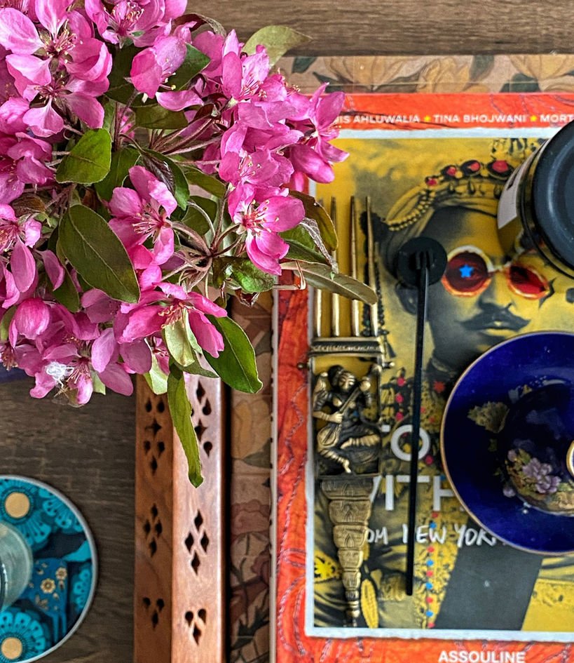 An overhead flat lay: hot-pink crabapple blossom, a saffron art book, a cobalt bowl, a turquoise enamel plate, a small brass figure and a carved wooden incense tower on dark walnut.