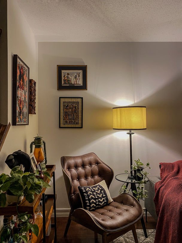 A brown tufted leather chair under an amber floor lamp, with a wall of framed folk paintings and a red throw.
