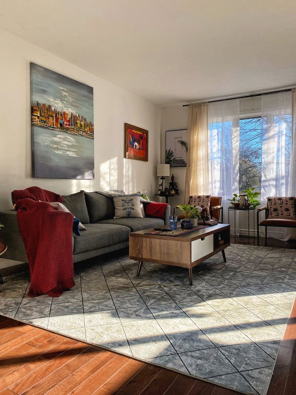 A grey sectional with a deep red throw, a walnut coffee table and a cityscape canvas above, on a patterned rug.