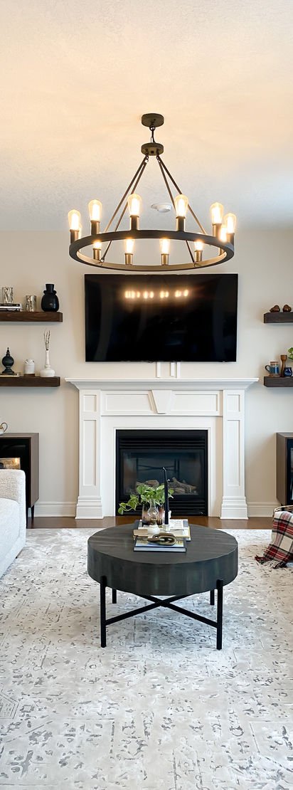 A white fireplace with a television above it, a black orb chandelier, a round black coffee table and a terrazzo-pattern rug.